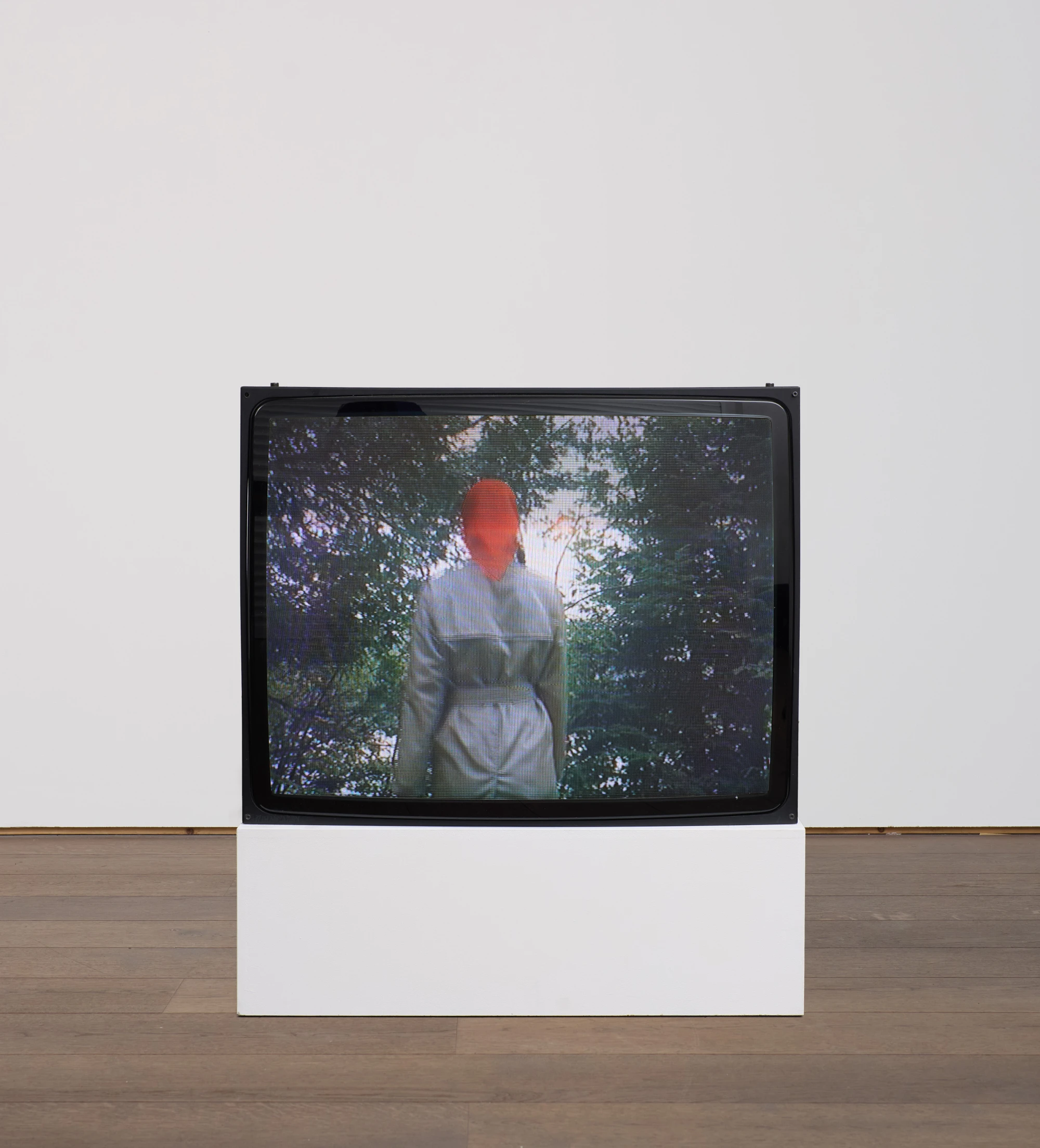 A black block monitor on the ground of a white-walled space. On the screen is a figure wearing a beige trench coat and red headscarf, with her back turned, walking toward an orange sun.